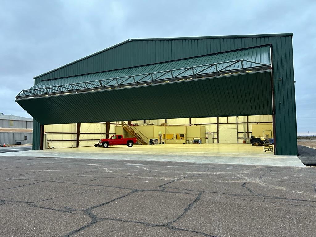 Large green industrial hangar with an open bay, containing a red pickup truck, some industrial equipment, and a yellow interior structure, under a cloudy sky.