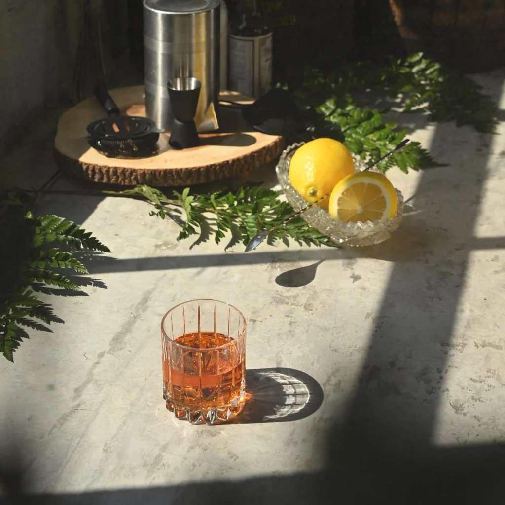 Hotel Tango Sazerac cocktail on a gray surface with lemons and bar tools in the background.