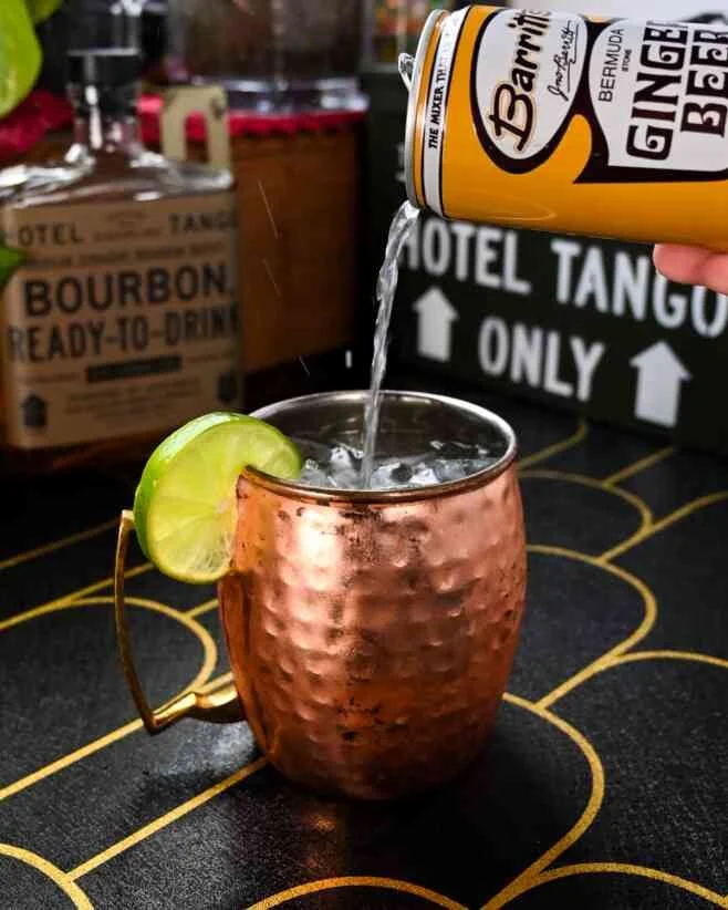 A Hotel Tango bourbon mule in a copper mug with lime garnish being filled with ginger beer from a can, on a black and gold table in a bar or restaurant setting.