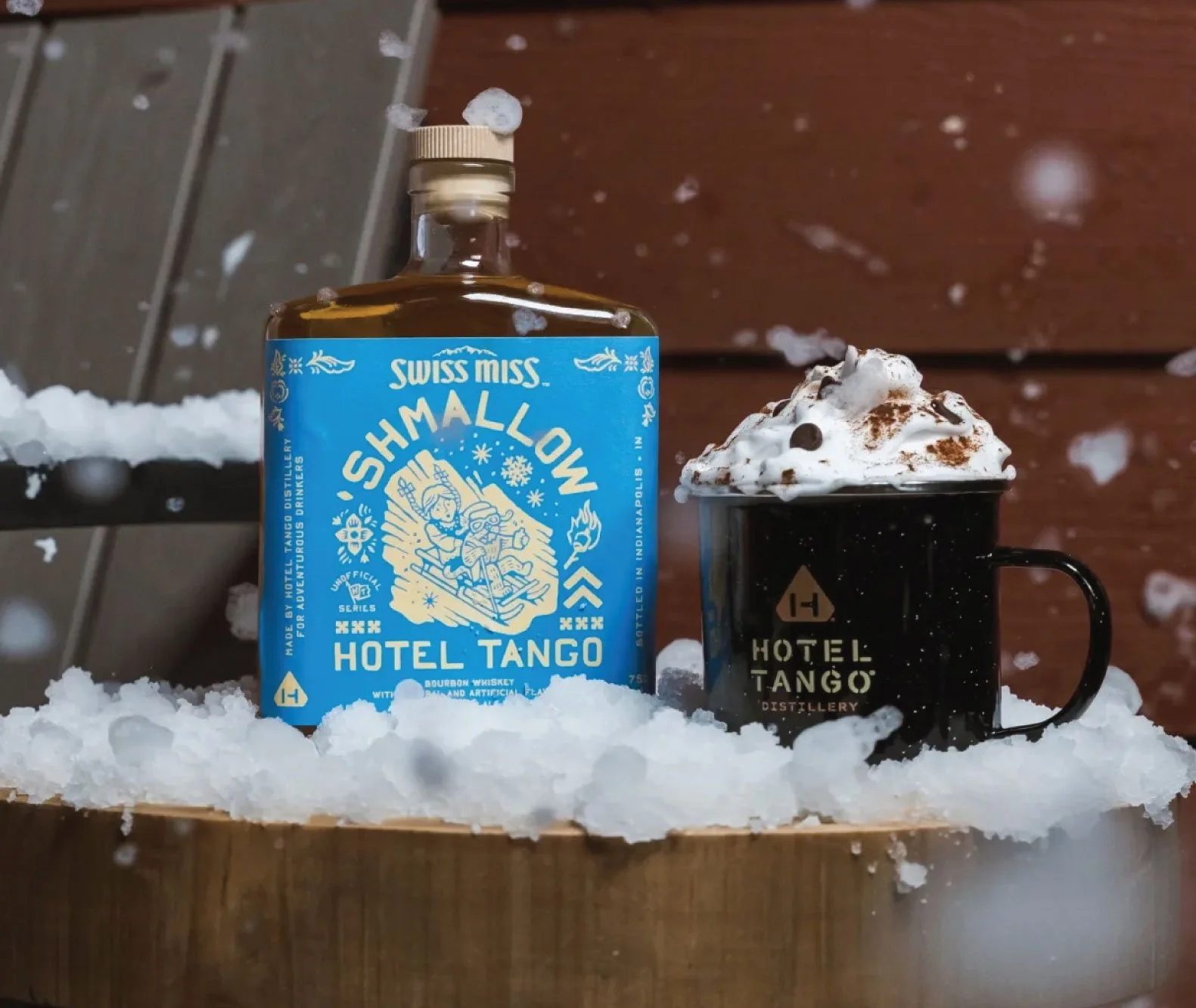 Hotel Tango Swiss Miss Shmallow Whiskey bottle with a mug of hot chocolate in a snowy setting.