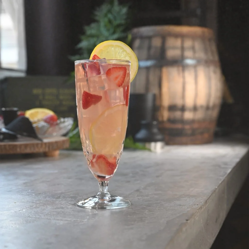 A Hotel Tango Strawberry Lemon Shakeup in a tall glass of lemonade infused with strawberries and lemon slices, garnished with a lemon wedge, on a bar or table surface with a rustic wooden barrel and greenery in the background.