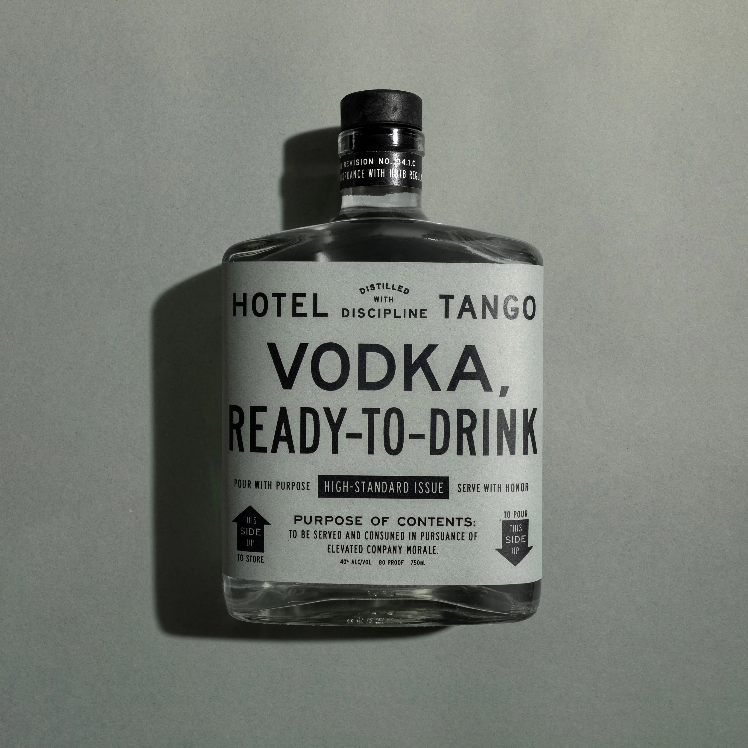Clear glass bottle of Hotel Tango vodka with a black cap, placed on a gray surface. The bottle's label features bold black and white text with details about the vodka.
