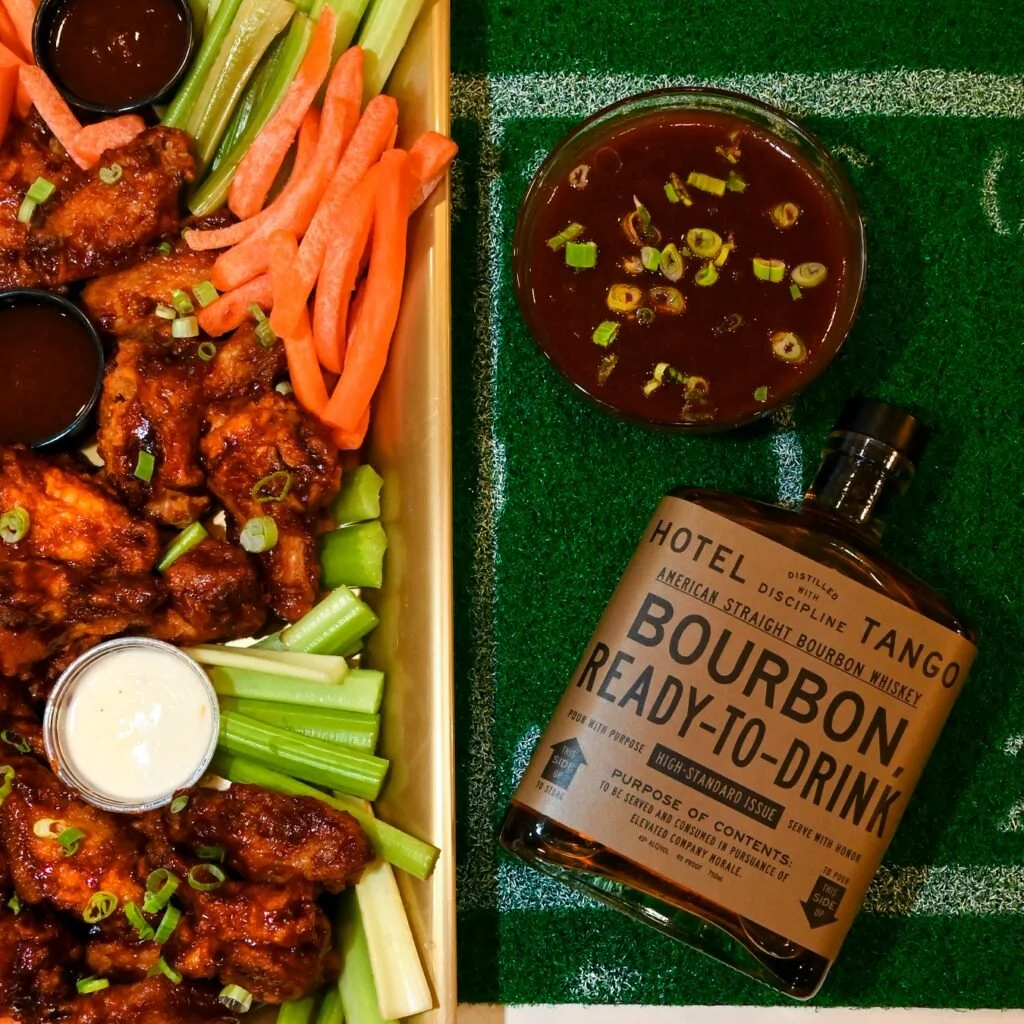 A Hotel Tango BBQ Bourbon chicken wing platter with barbecue sauce, celery sticks, carrot sticks, and dipping sauces, alongside a shot of bourbon ready-to-drink on a sports-themed table.