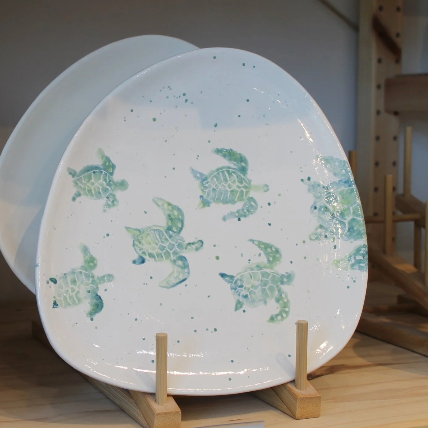 Decorative ceramic plate with a pattern of small green sea turtles and splatters, displayed on a wooden stand inside a shop or gallery.