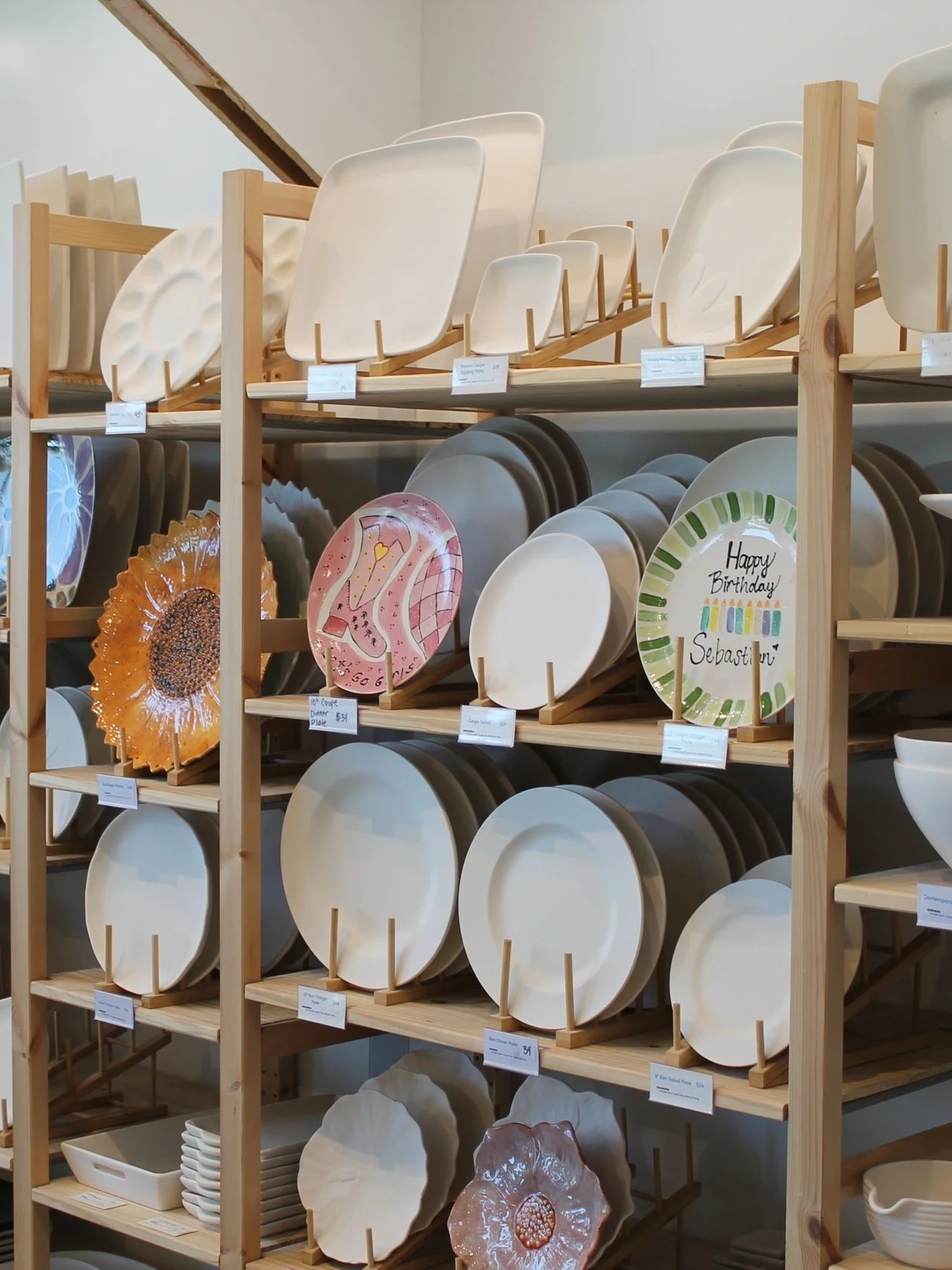 Display of decorative ceramic plates on wooden shelves in a store. Several plates feature colorful designs, including a birthday greeting and a pink pattern, with some resembling flowers or abstract art.