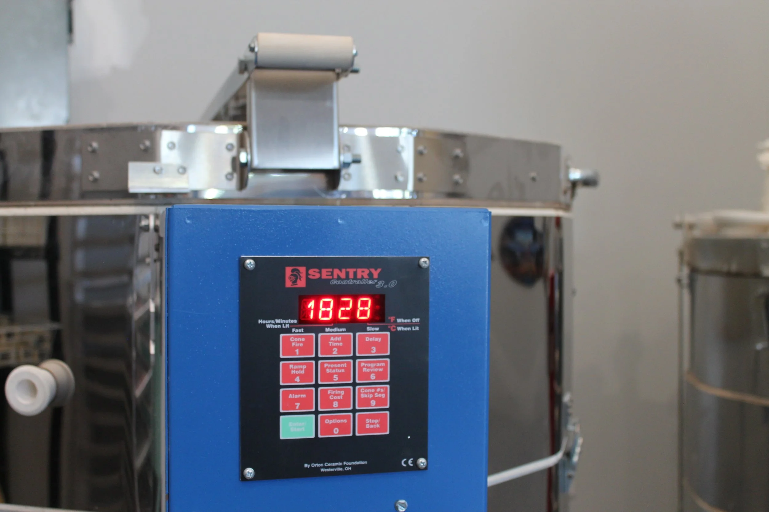 Close-up of a blue temperature control device with a digital display showing "1828" and various buttons labeled with functions like "Cone Fire," "Add Time," and "Options." In the background, there is a metallic piece of equipment with a roller.