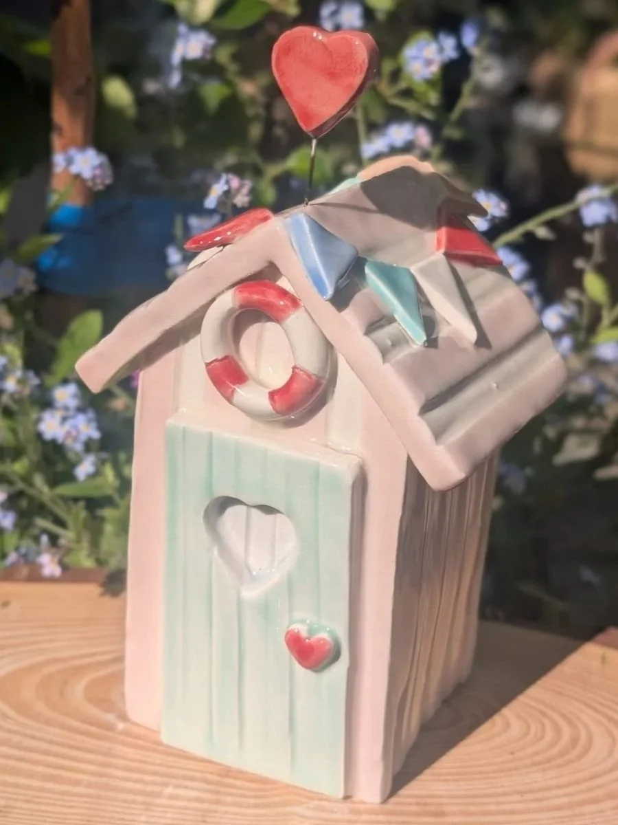 Decorative ceramic house with colorful heart-shaped and leaf-shaped accents, set outdoors against green foliage and small flowers.