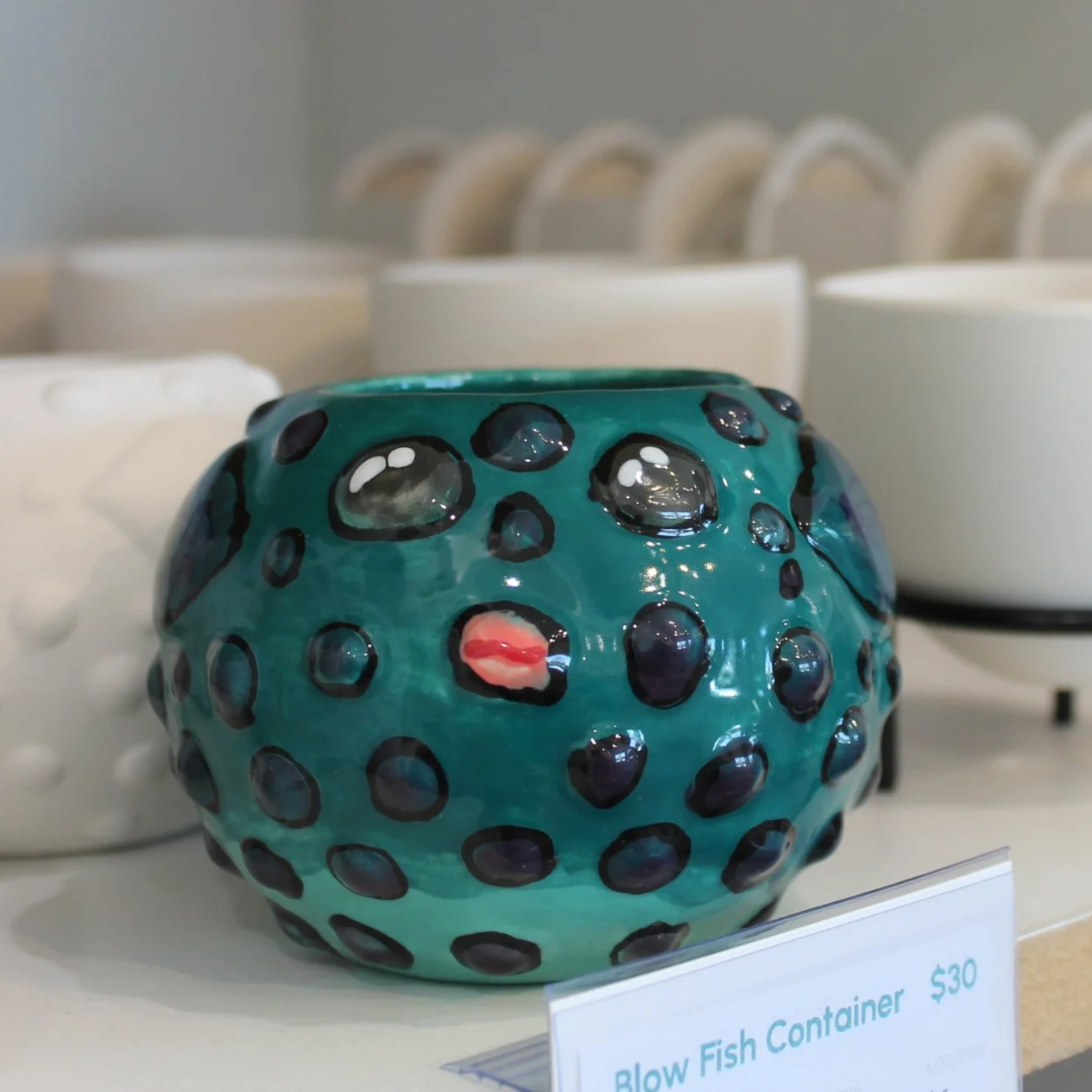 A round, ceramic blow fish container in turquoise color with black polka dots and a small red tongue, displayed on a store shelf with a price tag of $30.