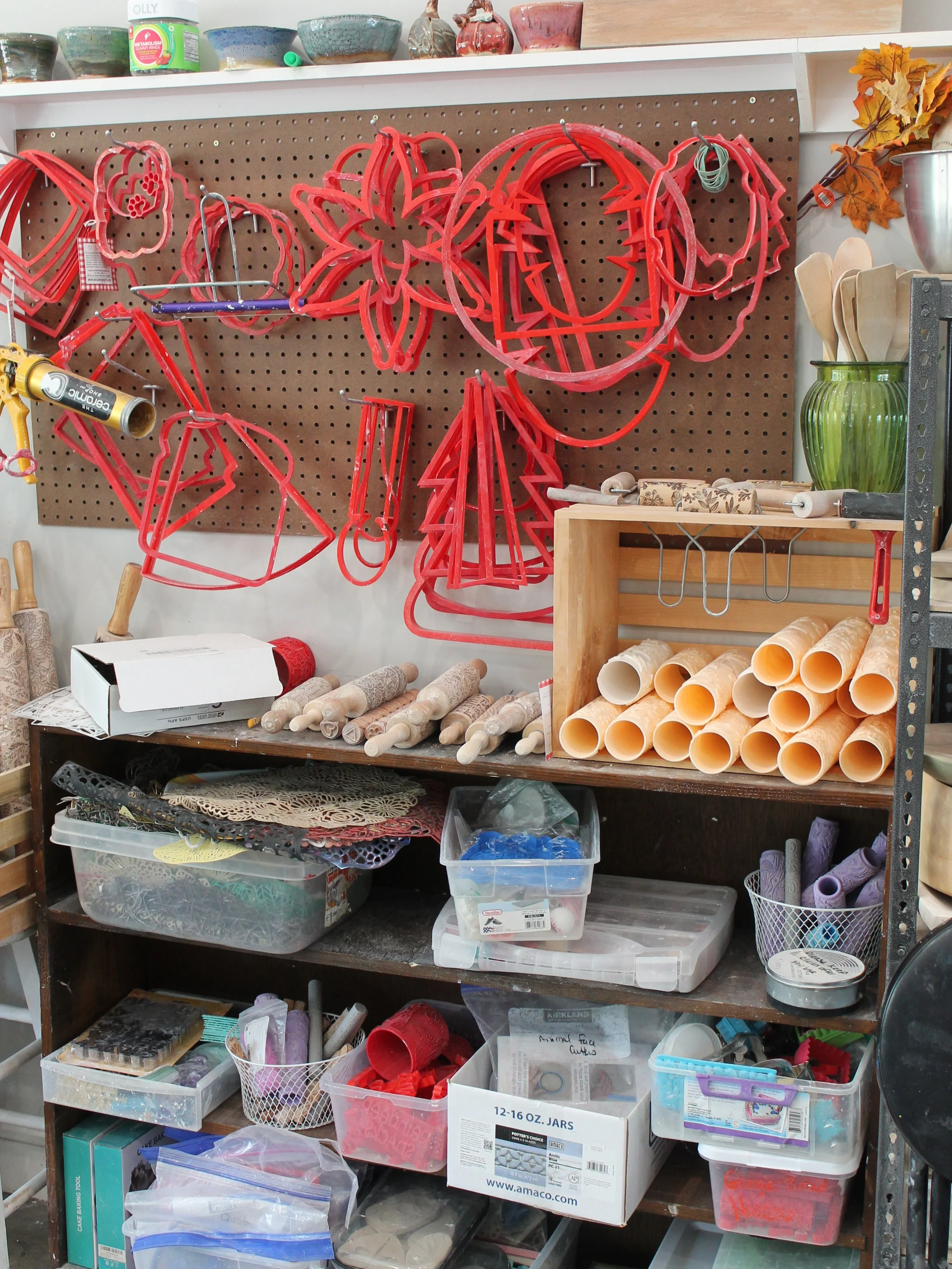 A workshop or craft supplies storage area with various items on shelves. Red plastic cookie cutters shaped like animals and holiday symbols hanging on a pegboard. Clear plastic bins filled with craft materials like rollers, foam sheets, and other tools. White boxes with storage labels.