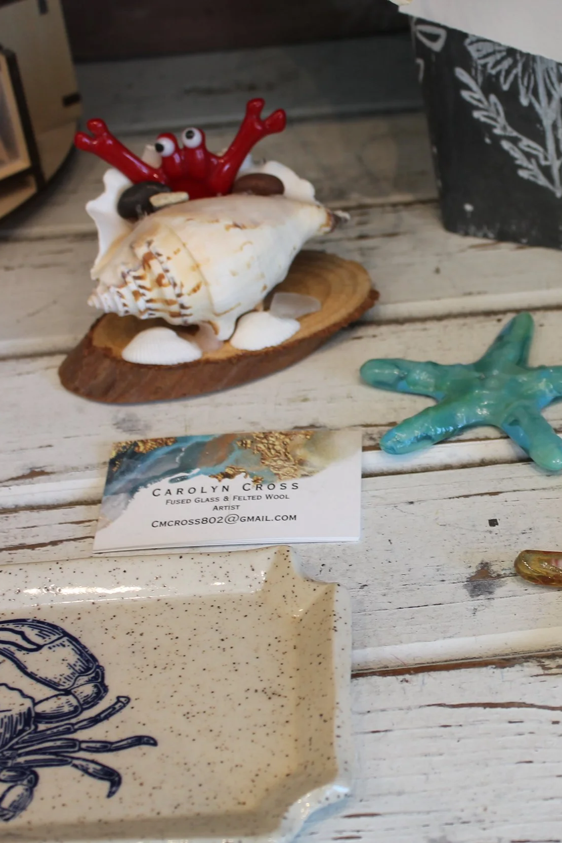 Decorative coastal-themed art pieces on a weathered white wooden surface, including a shell with a red crab figurine on top, small ceramic starfish, and a business card for artist Carolyn Cross.