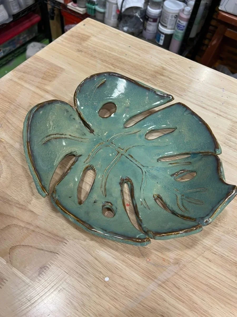 A ceramic dish in the shape of a monstera leaf, glazed in shades of green and brown, placed on a wooden surface with art supplies in the background.