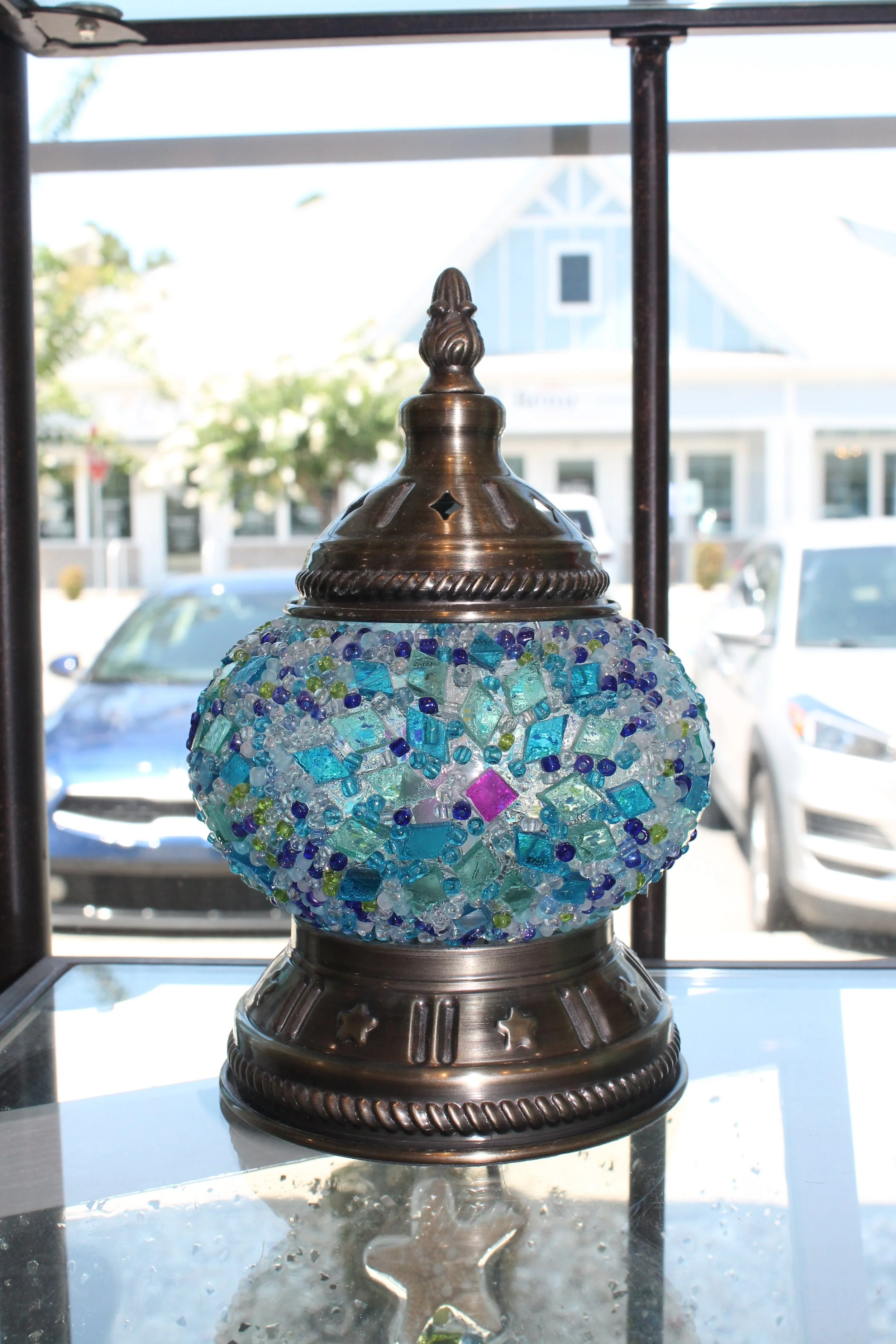 Close-up view of a decorative mosaic glass lamp with a metal top, set on a glass surface with reflections, with a blurred background of parked cars and buildings outside a window.