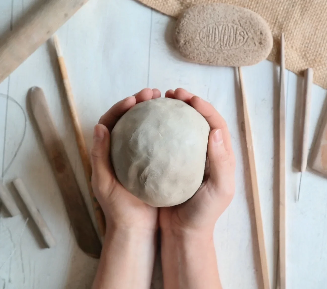 Hand holding a smooth, rounded stone on a table with various clay sculpting tools and a piece of burlap, and a stamped rock that says 'CINDOS' in the background.