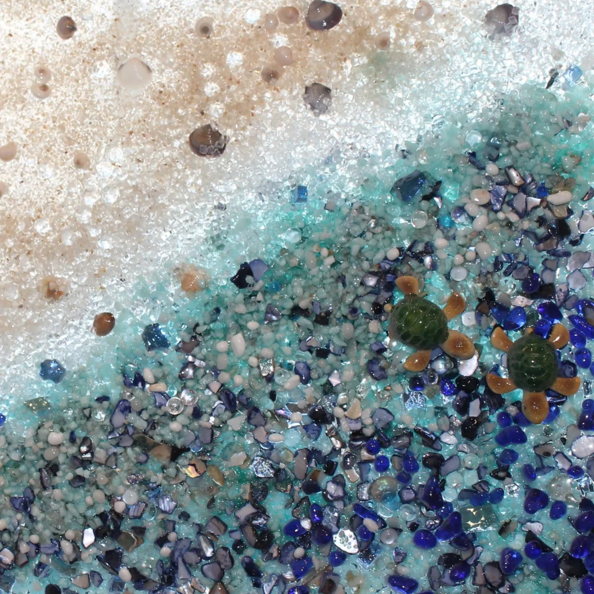 Two small green turtles with orange flippers swimming among colorful glass and shell fragments in shallow water.