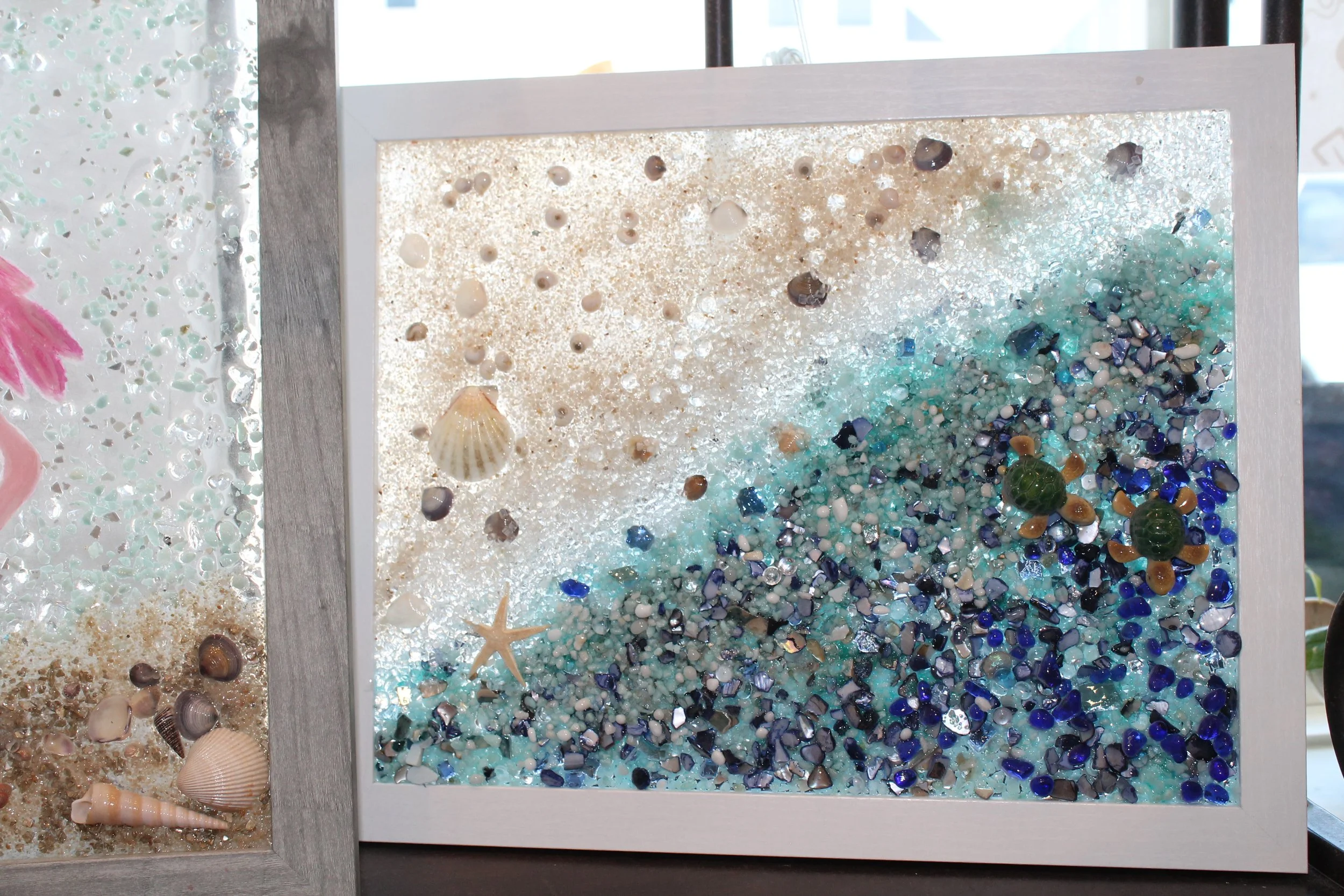 Two shadow boxes filled with various seashells and small decorative glass pieces, one with a white frame and the other with a silver frame, displayed near a window.