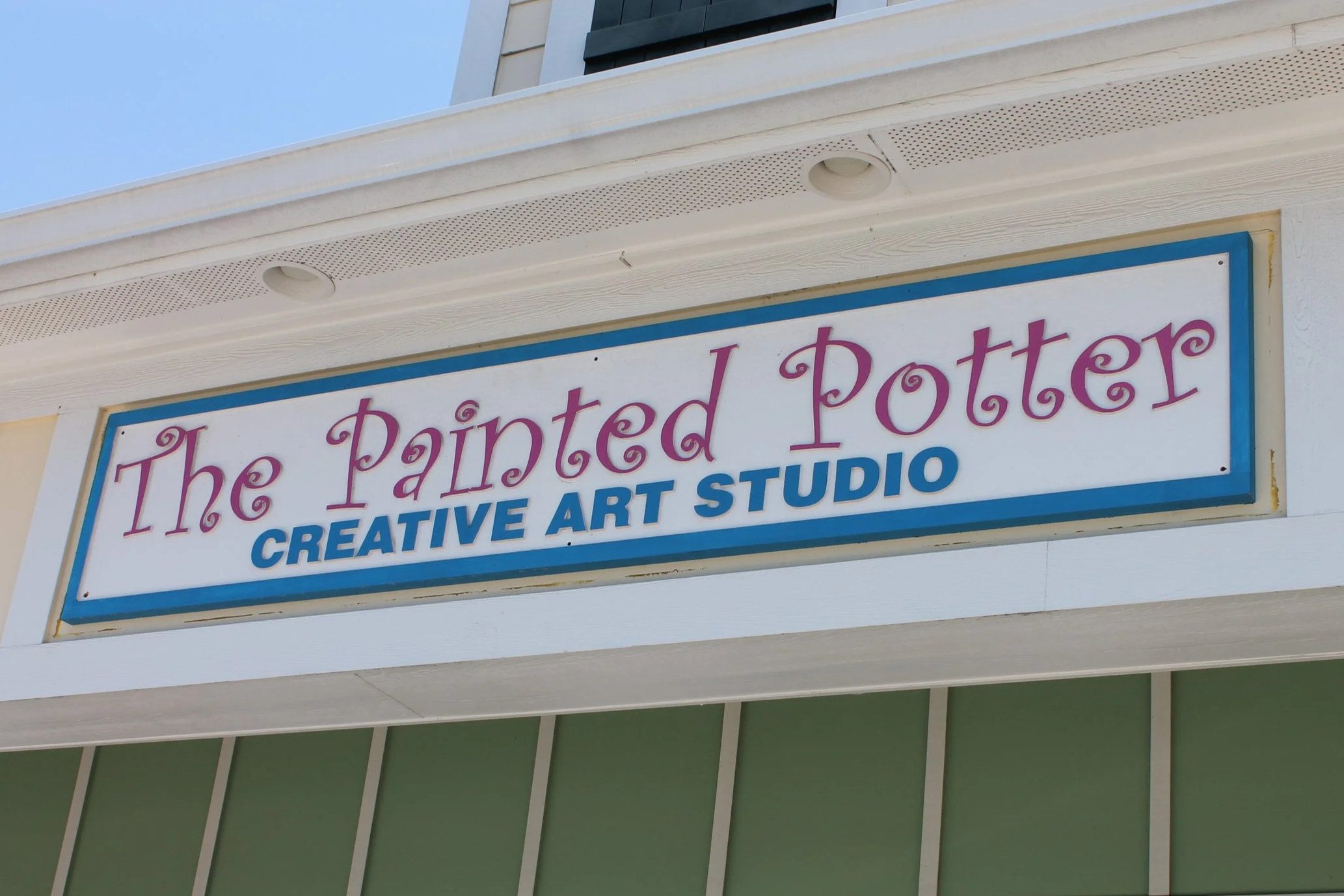 Sign on the wall of a building reading 'The Painted Potter Creative Art Studio' with a blue border around the sign, part of the building's exterior wall, with a clear blue sky above.