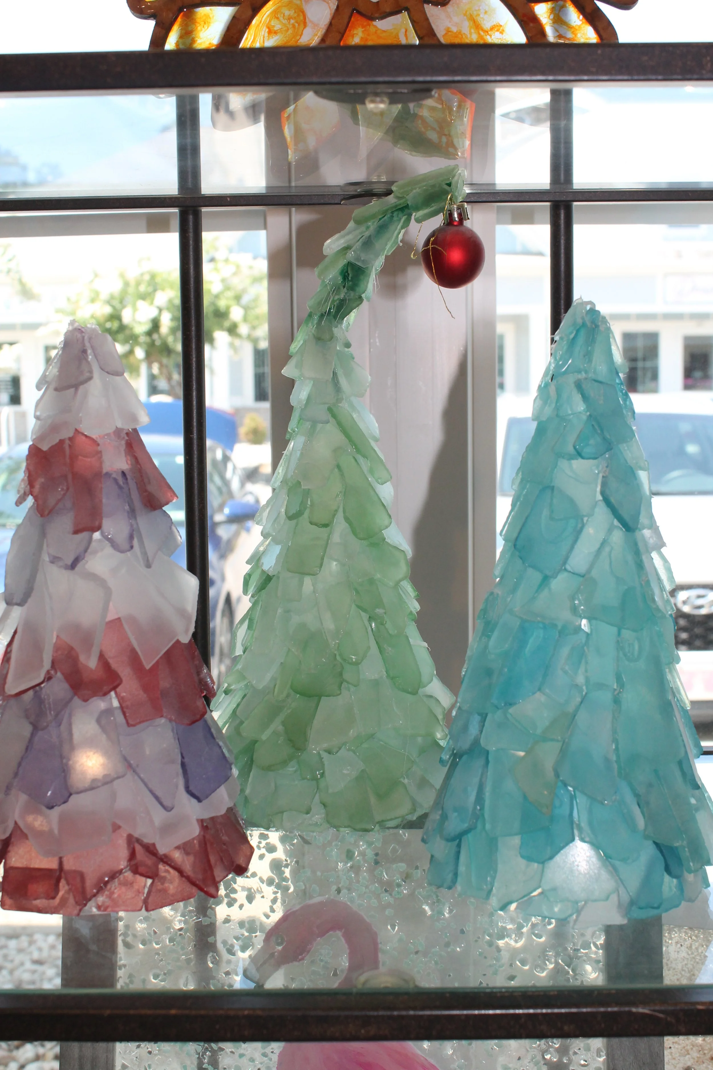 Decorative glass Christmas trees in red, green, and blue, displayed on a window ledge with a painted flamingo in front, overlooking a sunny outdoor scene with parked cars and buildings.