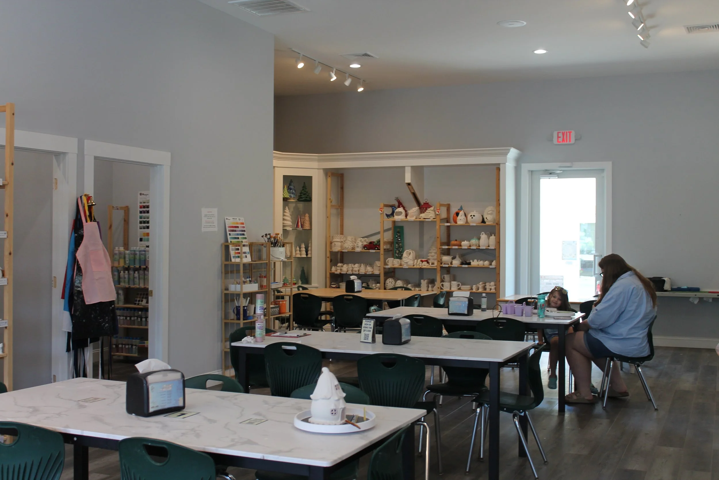 Interior of a craft or pottery store with shelves filled with ceramic decorative items, tables with chairs, and a woman with a child sitting at a table.
