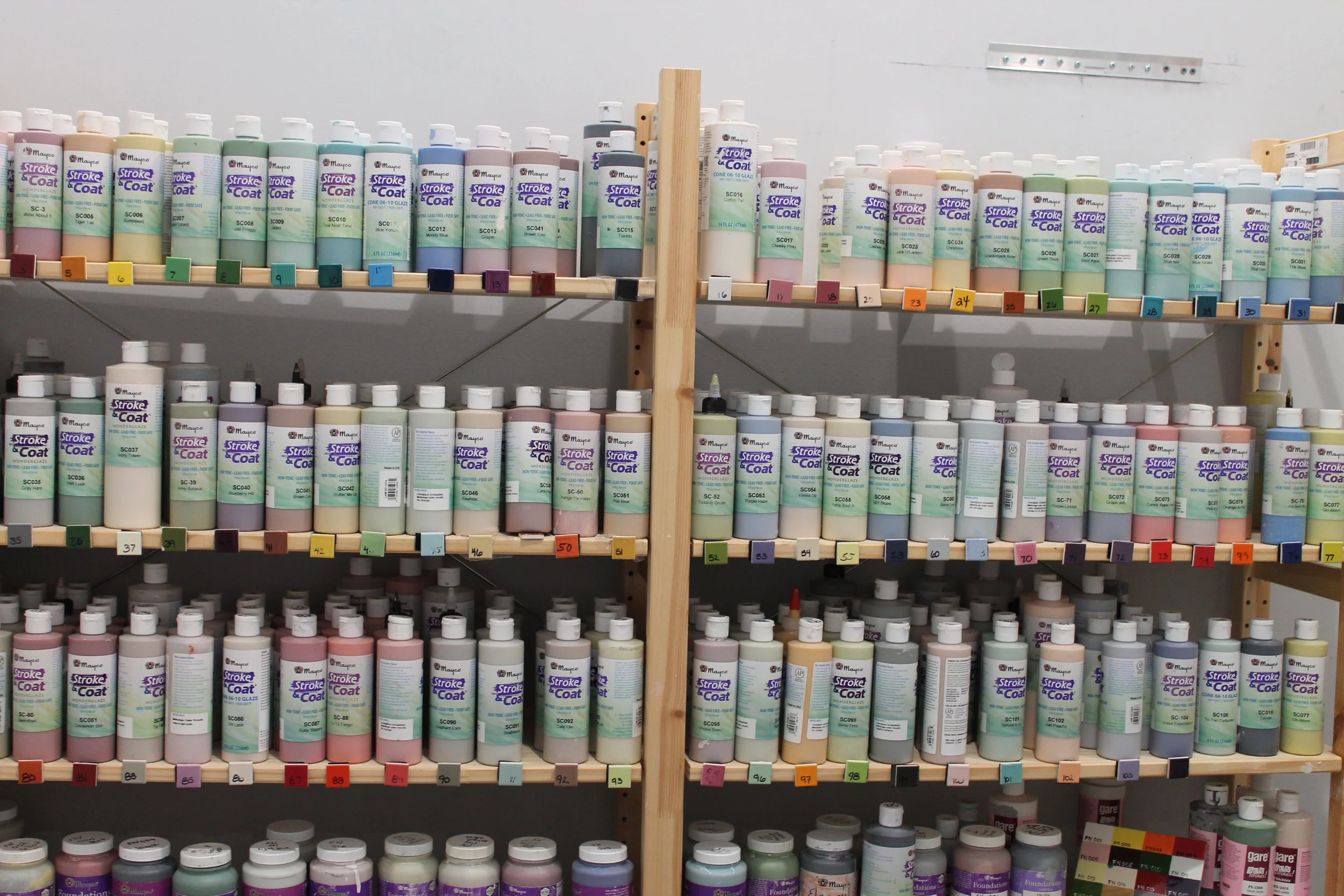 Various bottles of Mayco Stroke & Coating color paints arranged on wooden shelves, organized by color and code.
