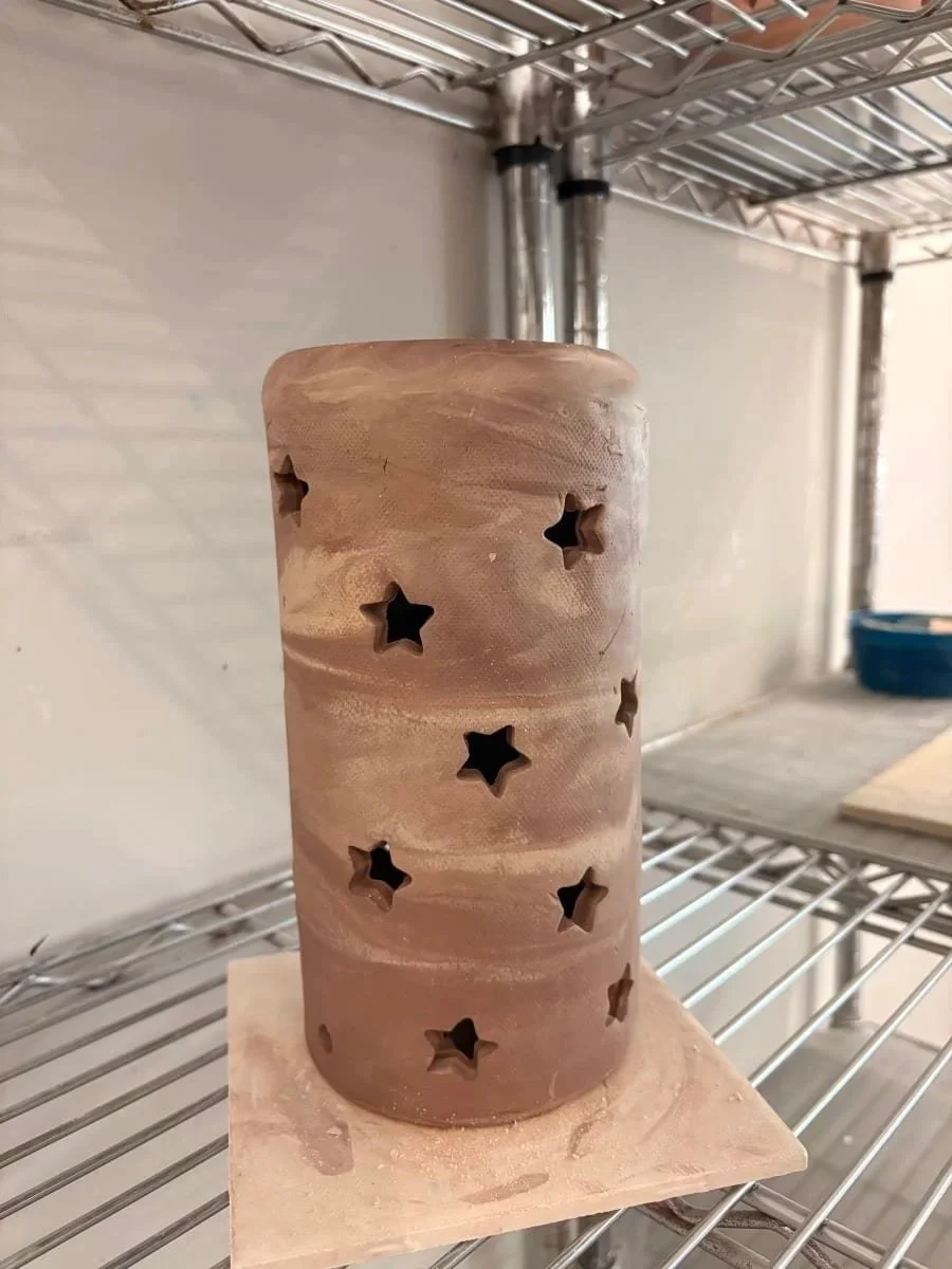 A cylindrical ceramic object with star-shaped cutouts, placed on a piece of cardboard on a metal wire shelf.