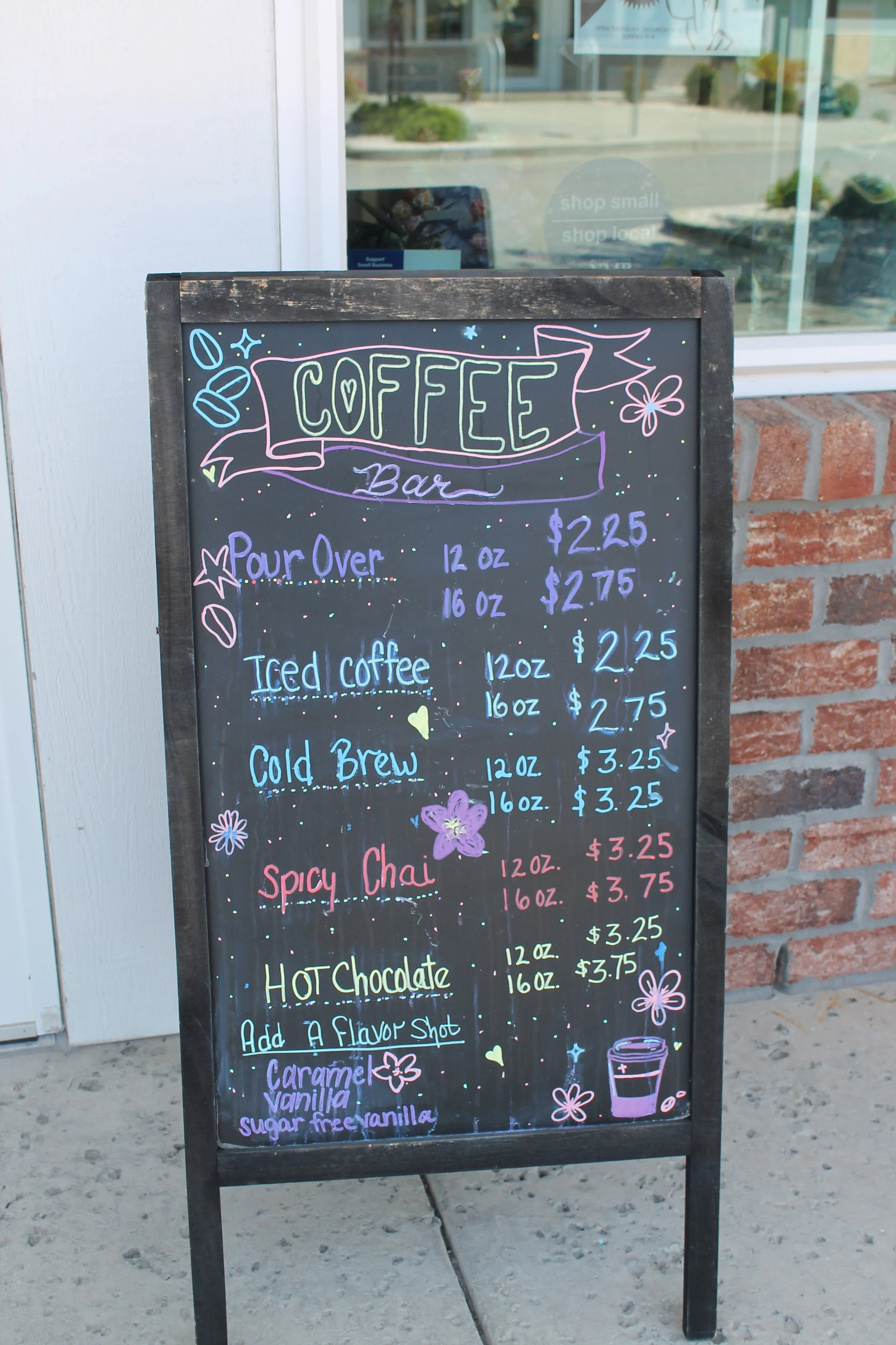 Colorful neon-style signboard outside a coffee bar listing various coffee drinks and prices, decorated with doodles of flowers, stars, and coffee beans.