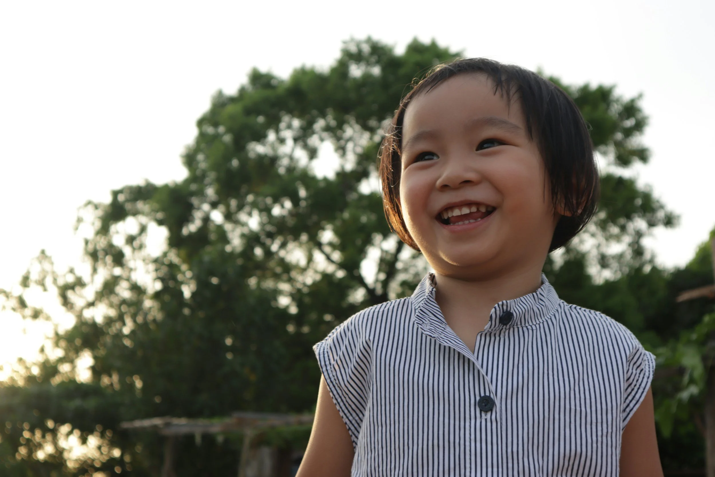 A young boy with short dark hair smiling outdoors with trees and a bright sky in the background.