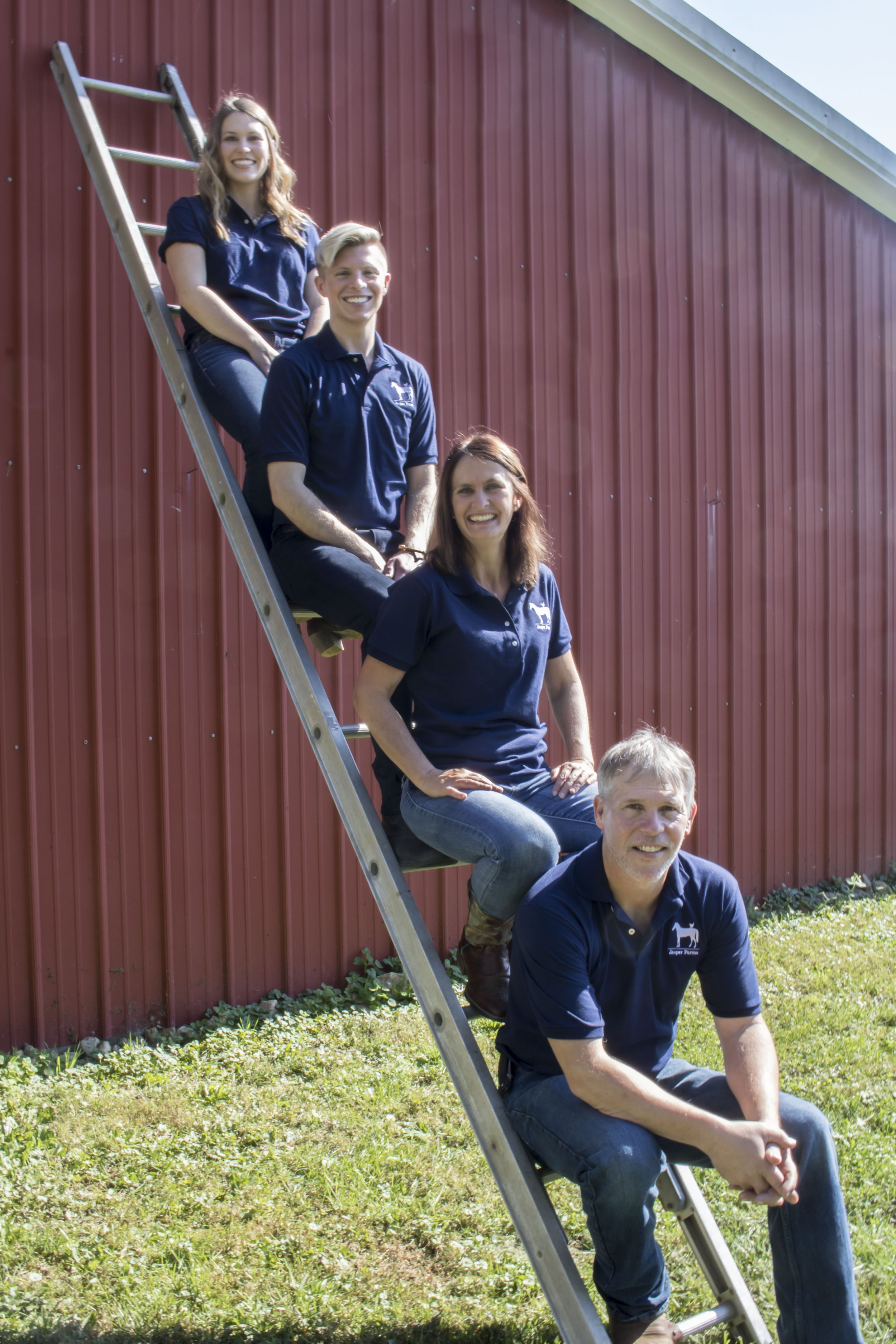 Four people, dressed in matching navy shirts with a white logo, are posing on and around a ladder outside a red building with vertical siding. They are smiling and appear to be part of a team or group.