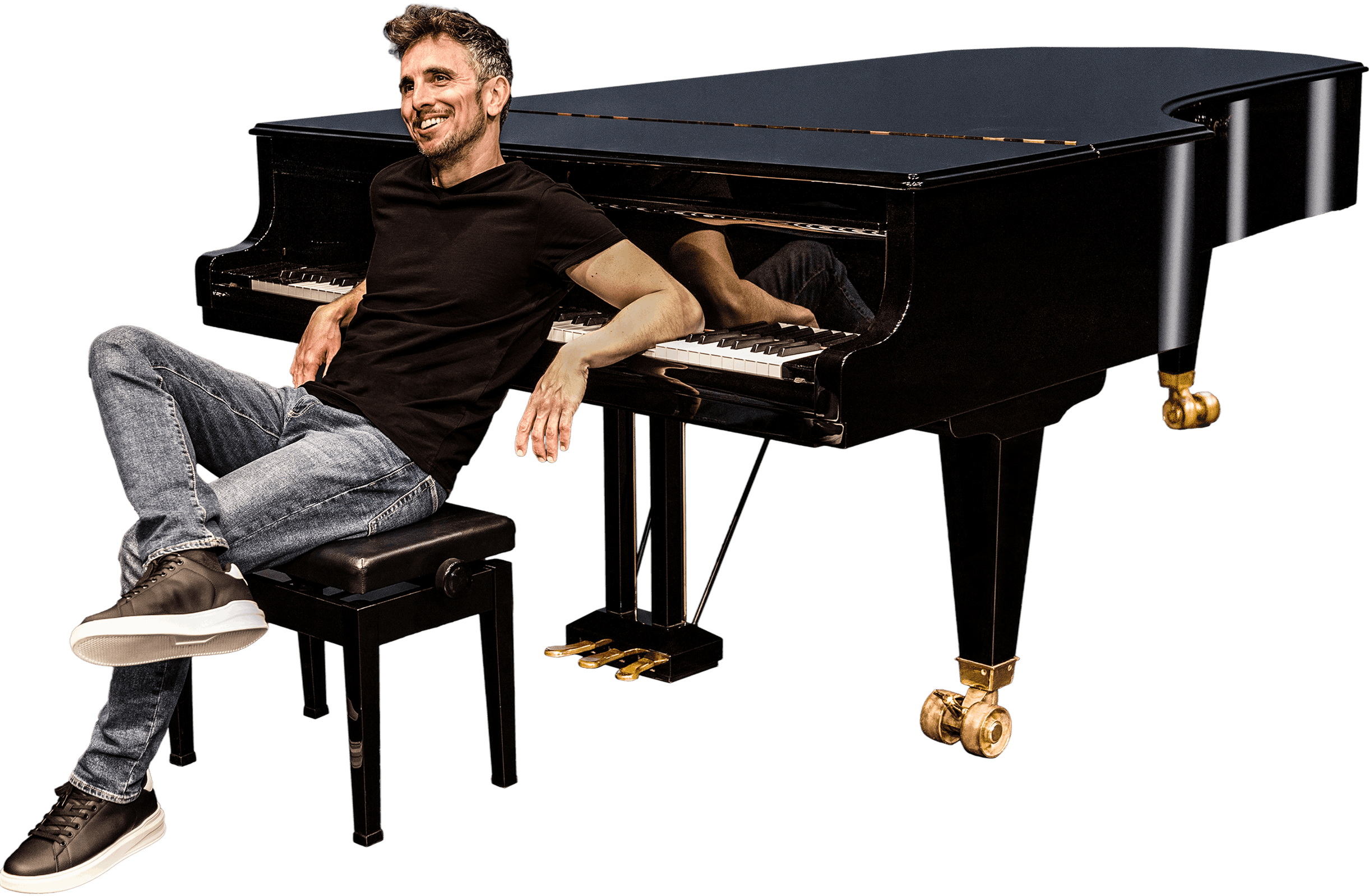 A man in a black t-shirt and jeans sitting on a piano bench smiling, with one arm resting on the open black grand piano next to him, against a black background.