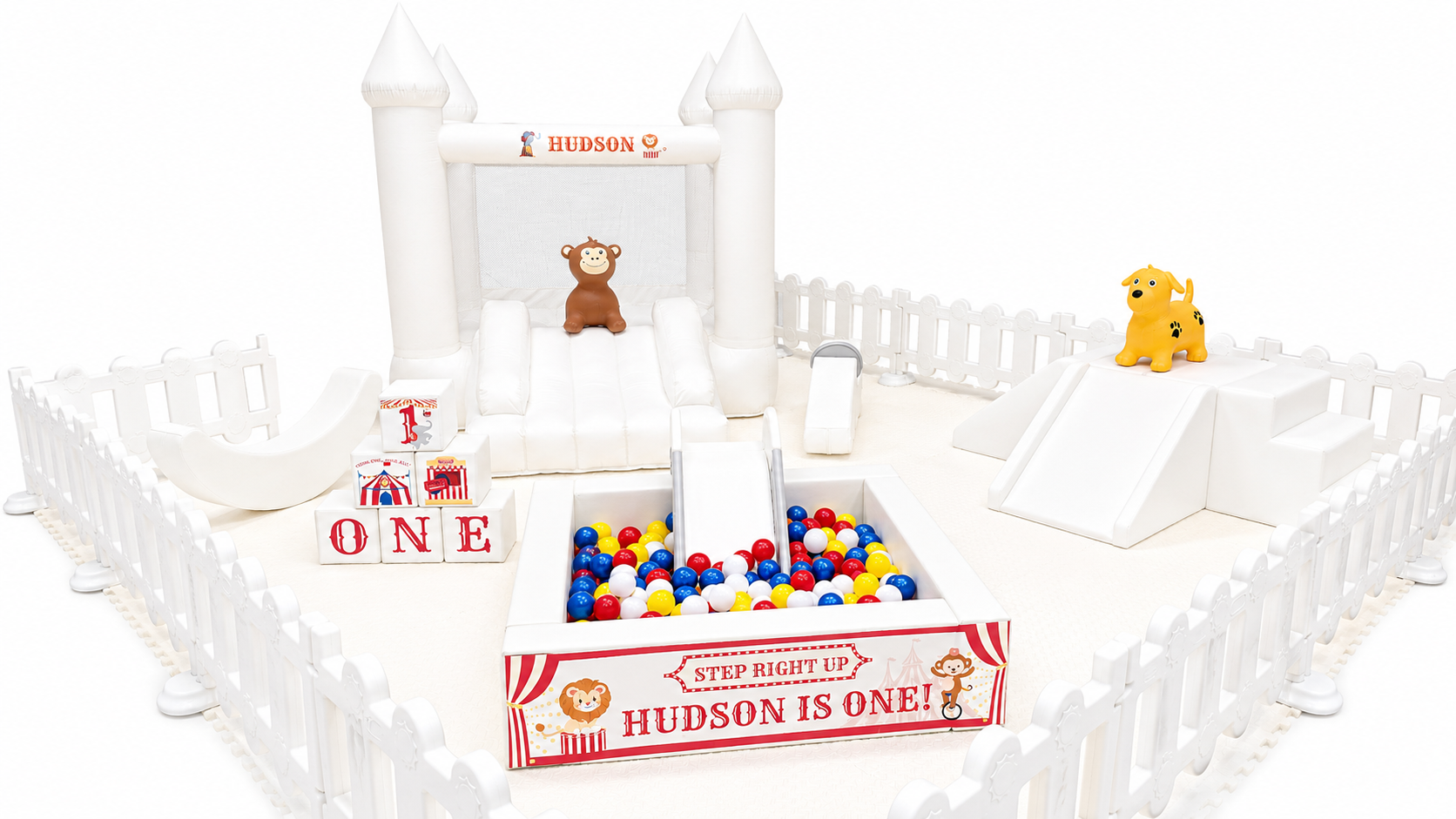 White birthday party bounce house with a slide, surrounded by white picket fence. Decorations and toys include blocks spelling 'ONE', stuffed animals of a bear, a dog, and a lion, and a ball pit with red, blue, white, and yellow balls. Sign reads 'Step Right Up Hudson is One!'