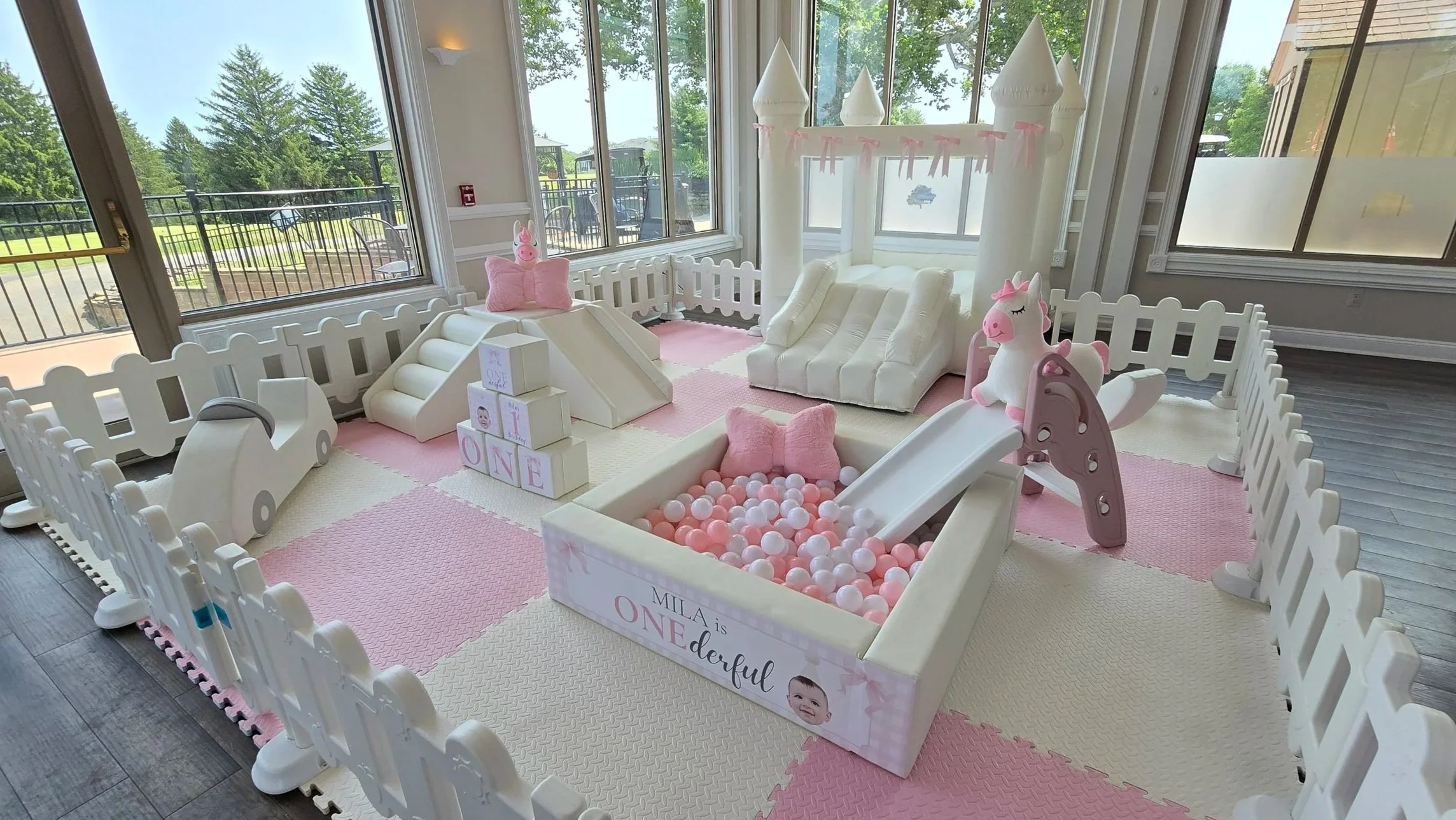 Indoor princess-themed birthday party setup with a white castle playhouse, pink and white decorations, a ball pit with pink and white balls, and a fenced area with pink pillows and a slide.