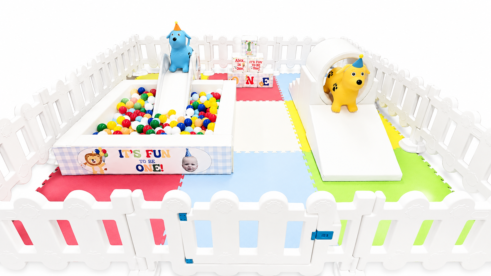 A children's indoor play area with a small ball pit, two colorful dog-shaped ride-on toys, and a foam mat with blocks and a birthday message for a first birthday celebration.