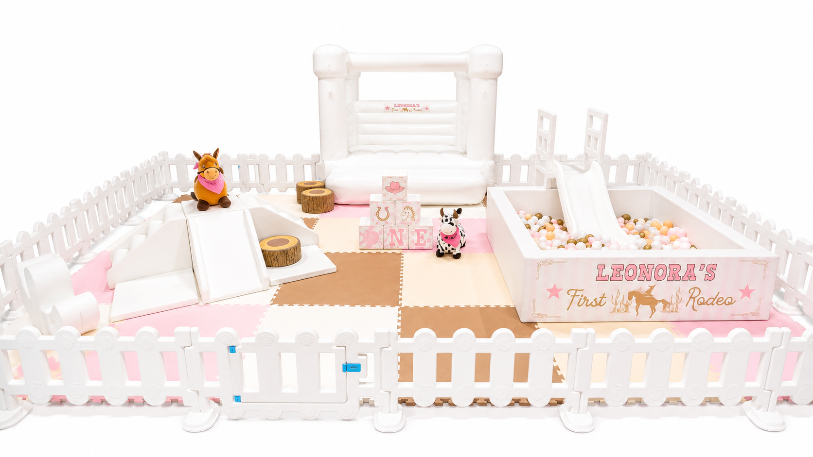 A miniature play area themed for a child's first rodeo. It features a white picket fence, a small slide into a ball pit, a dollhouse-like structure, plush animals, and decorative blocks spelling 'N E' with a horse and cow pattern.