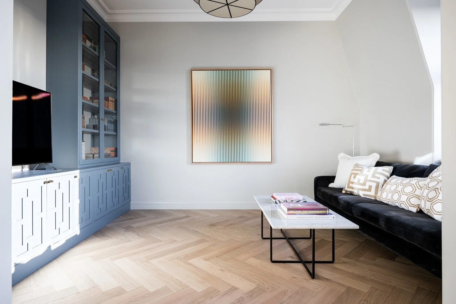 Lys stue med hvite vegger, svart sofa med puter, kommode og bokhylle i blått, abstrakt kunstverk på veggen, gulv i lys tregulv, liten bord med bøker.