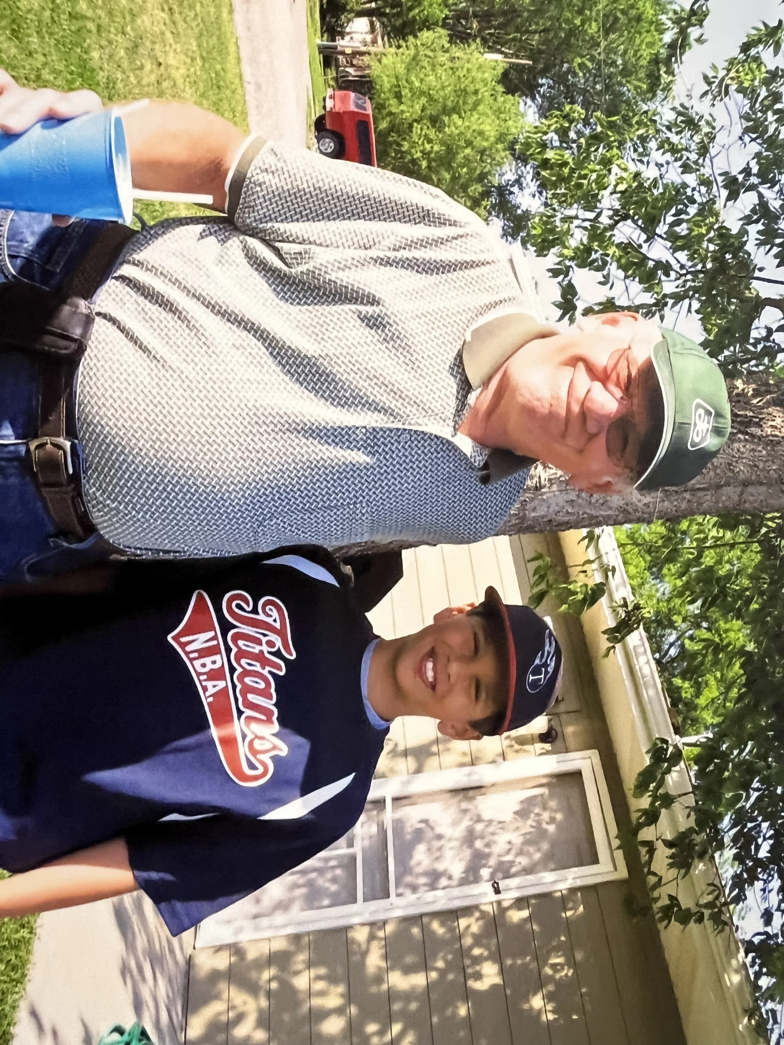 A man with glasses wearing a dark baseball cap and a Redskins sweatshirt taking a selfie outdoors in a garden area with trees and a house. A young boy is standing next to him, wearing a hat and glasses, smiling. There is a red toy truck in the background.