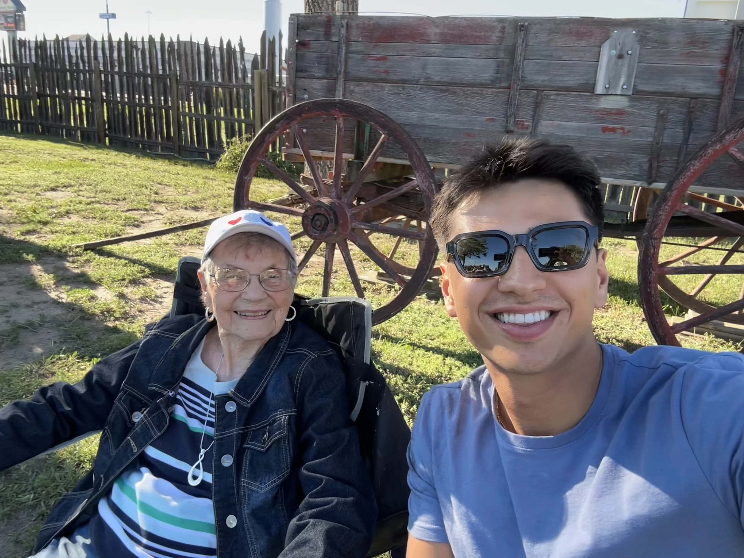 A selfie of an elderly woman with glasses and a white cap, and a young man wearing sunglasses, both smiling, outdoors near an old wooden wagon with red wheels.