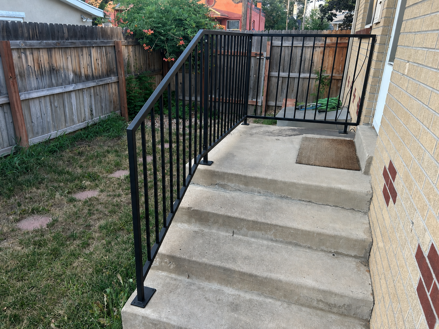 Concrete steps with a black metal railing outside a house, leading to a small porch area with a brown doormat, adjacent to a brick wall with a partially visible door, garden hose, and a grassy area with a wooden fence.