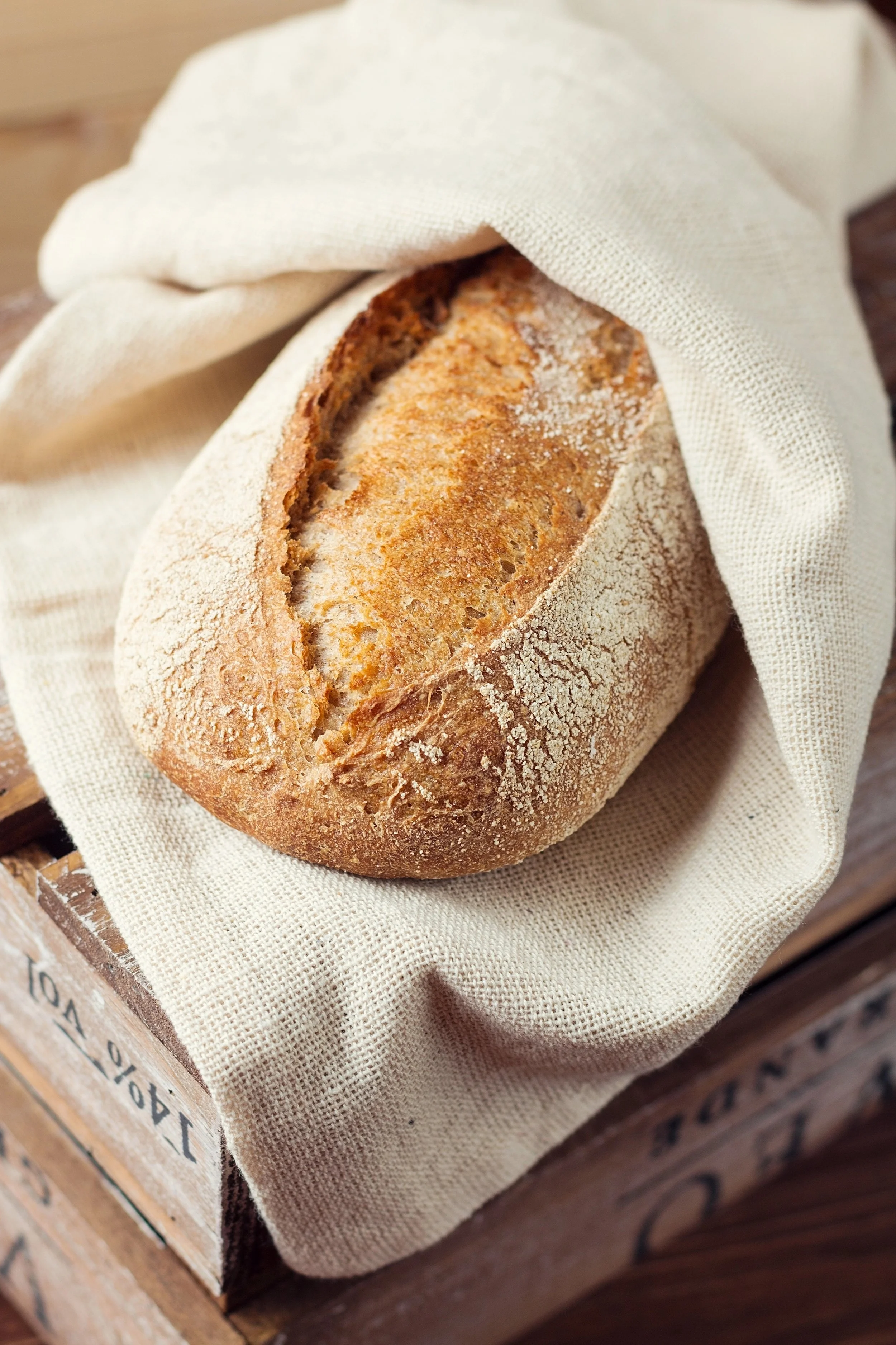 A loaf of bread with a crusty exterior resting on a beige cloth, placed on a wooden surface.
