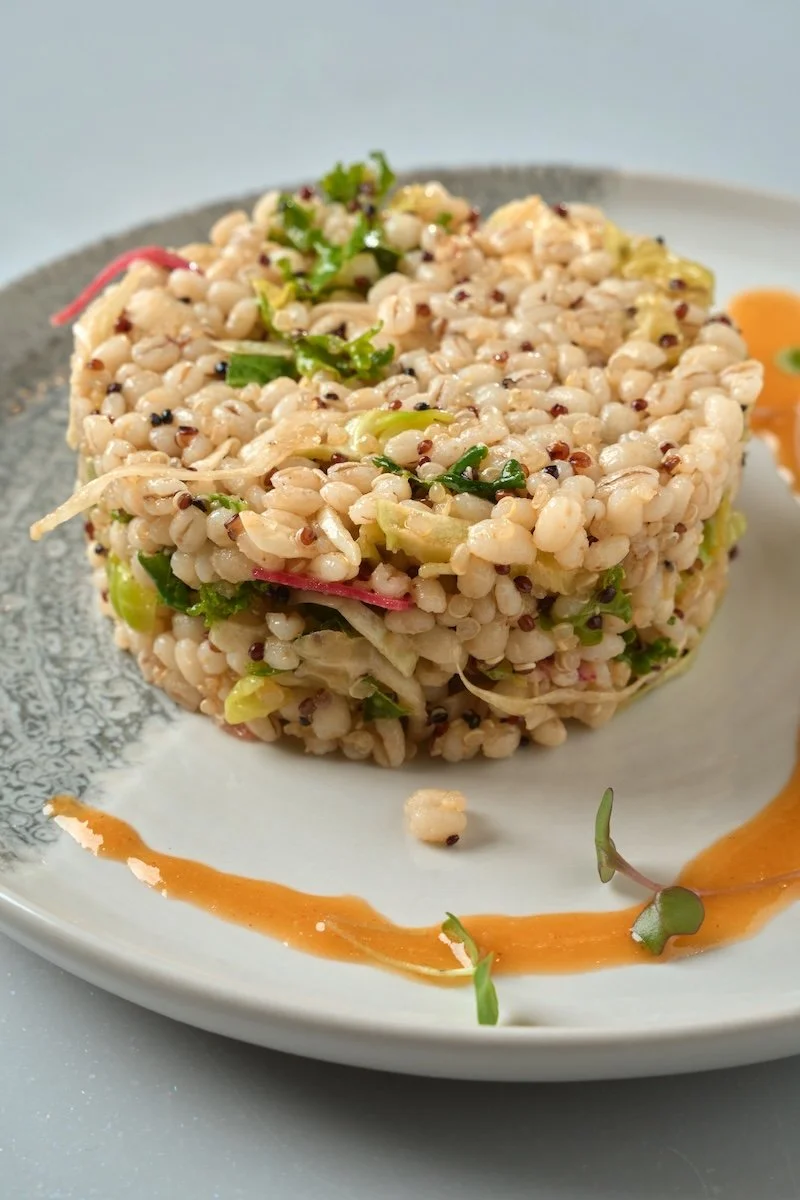A close-up of a round serving of barley salad garnished with microgreens on a plate, with a drizzle of orange sauce on the side.