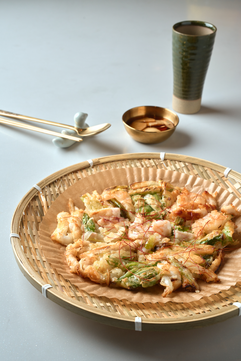 Fried tempura with green onions on a round bamboo tray, accompanied by a small bowl of dipping sauce, a green ceramic cup, and a white ceramic spoon with gold chopsticks on a light blue surface.