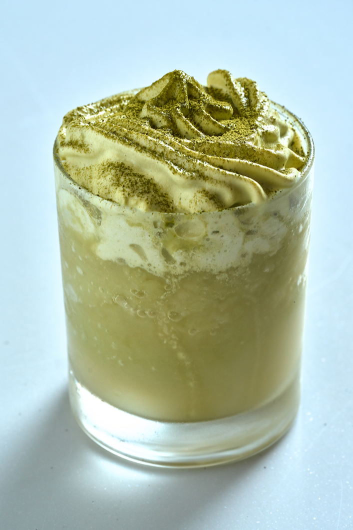 A green frozen matcha makgeolli with whipped cream and a dusting of matcha powder, styled in a clear glass against a soft background.
