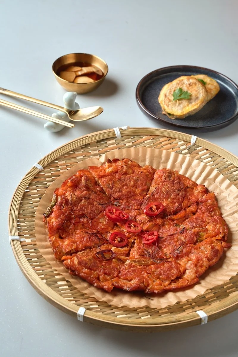 A traditional Korean kimchi pancake garnished with red chili peppers, served in a woven basket. Accompanying dishes include soy sauce in a gold bowl, a small dish with a fried egg topped with cilantro, and a pair of gold chopsticks and spoon.