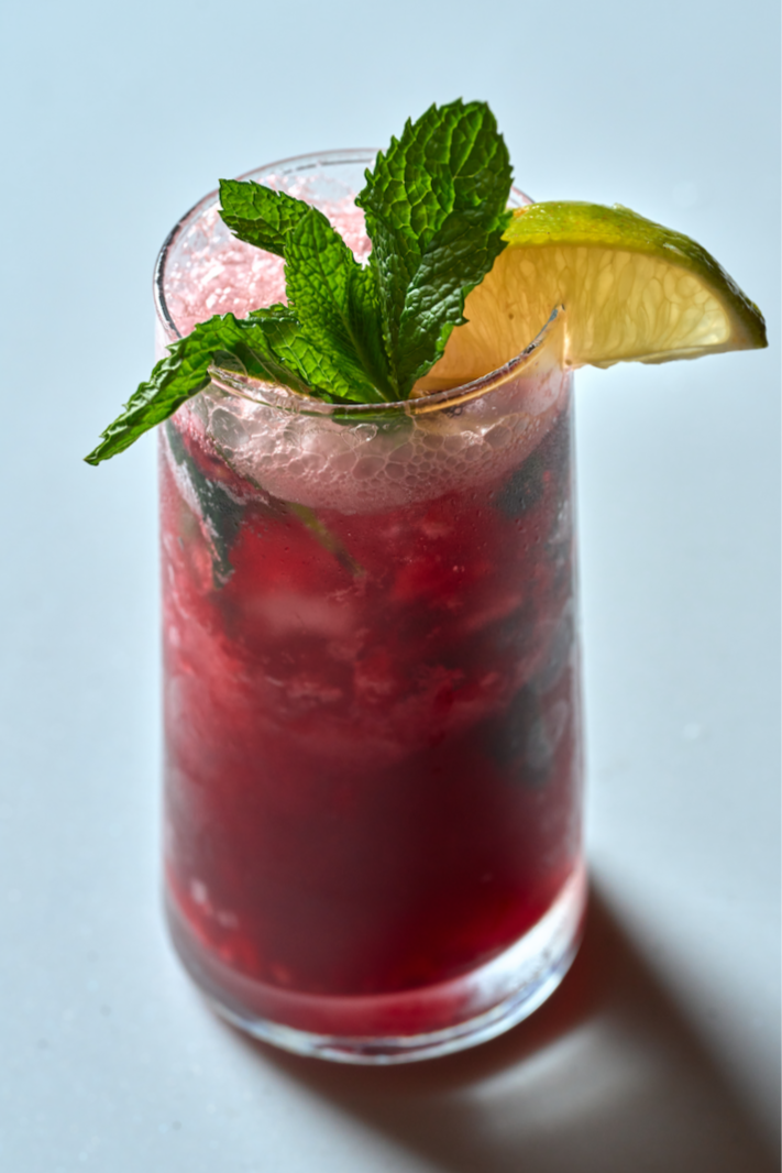 A deep red wildberry mojito served in a clear glass, garnished with fresh mint and a lime wedge, photographed against a light background.