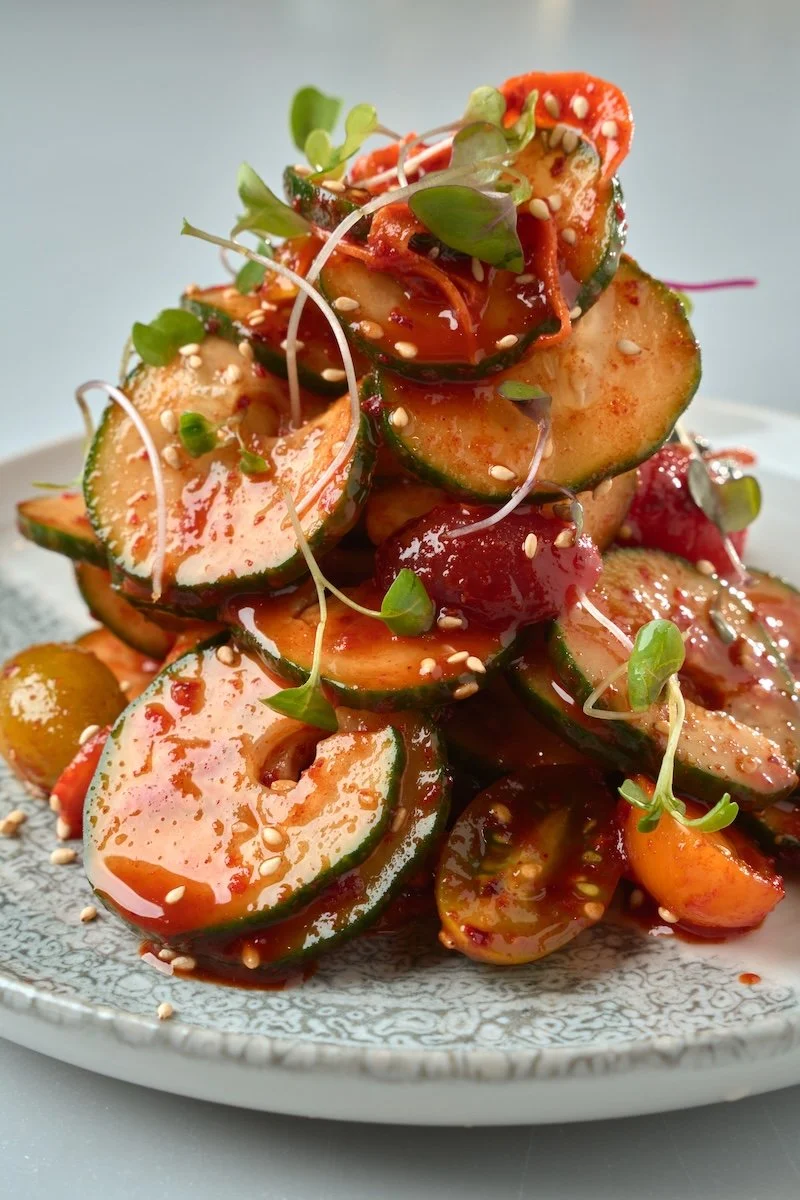 A plate of sliced cucumbers topped with sesame seeds and microgreens, dressed with a spicy red sauce.