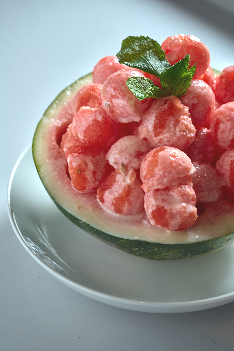 A halved watermelon filled with small watermelon balls and garnished with mint leaves, photographed on a clean, bright background.