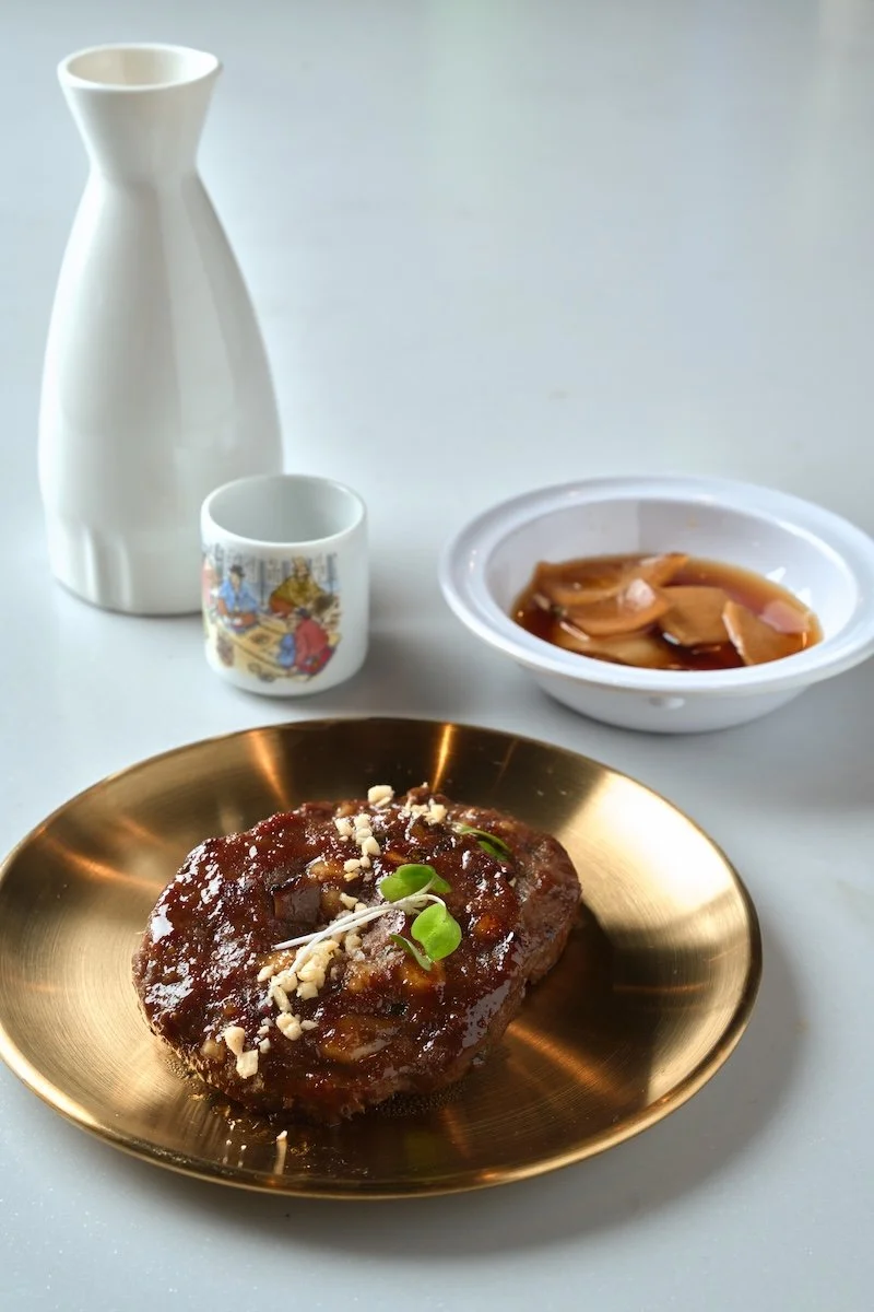 A plate with a large piece of glazed meat garnished with chopped nuts and microgreens, a bowl of broth with meat slices, a ceramic cup with a painted scene, a white vase, and a bowl of sauce or condiment on a white surface.