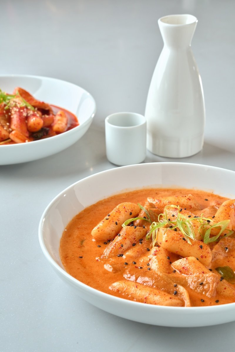 A bowl of spicy Korean tteokbokki with cylindrical rice cakes in a red sauce, garnished with green onions and sesame seeds, with side dishes and tableware in the background.