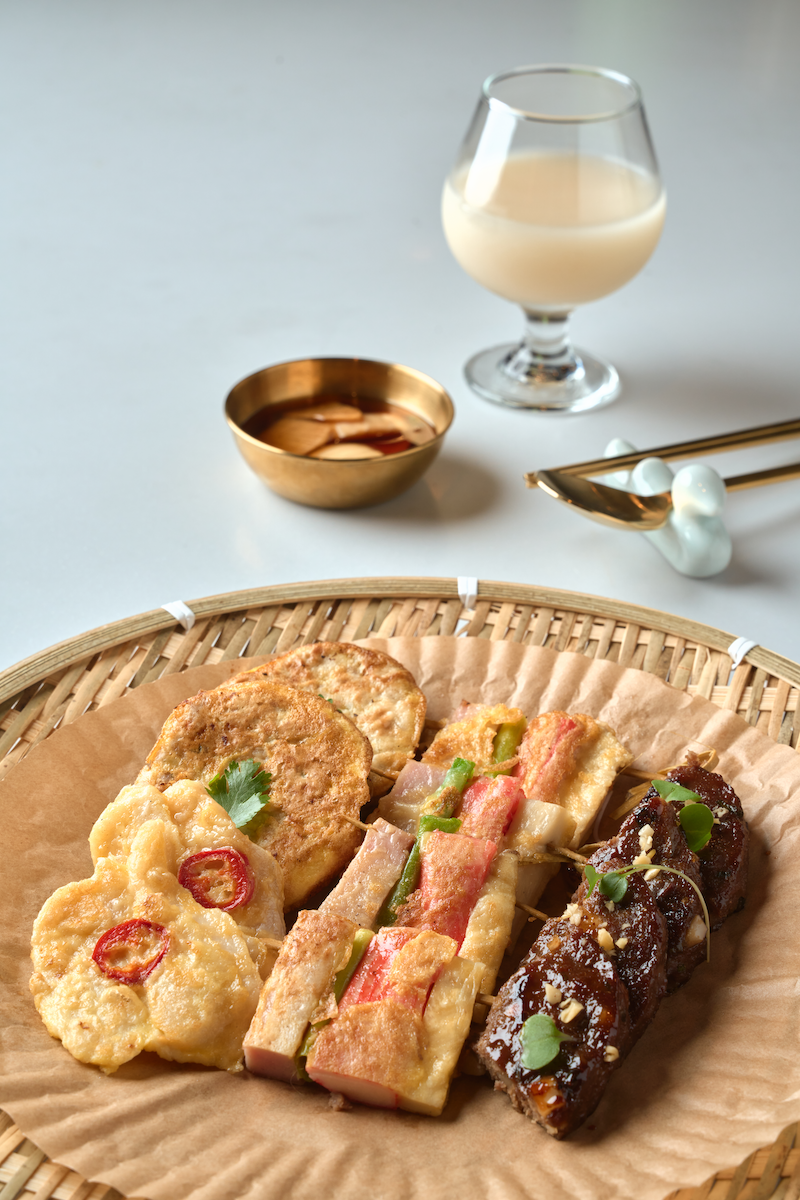 A round woven tray with assorted Asian-inspired appetizers, including fried potato patties, eggplant or tofu with chili slices, crab sticks with green onions, and glazed meat rolls garnished with microgreens, set on parchment paper. In the background, there is a glass of white wine, a small golden bowl, and gold-colored utensils on a white surface.