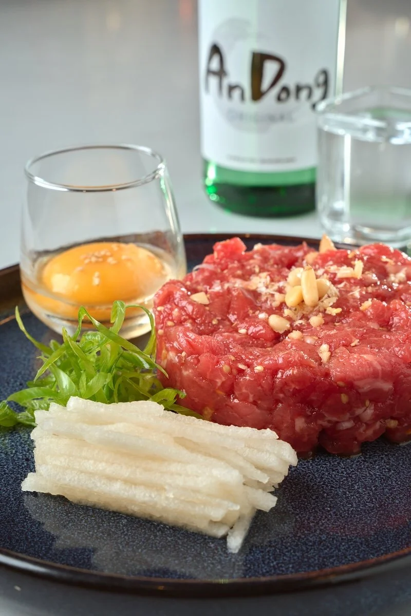 Raw beef tartare garnished with pine nuts on a black plate, accompanied by white bread sticks, fresh greens, a glass of raw egg, a small glass of water, and a bottle of Soju in the background.