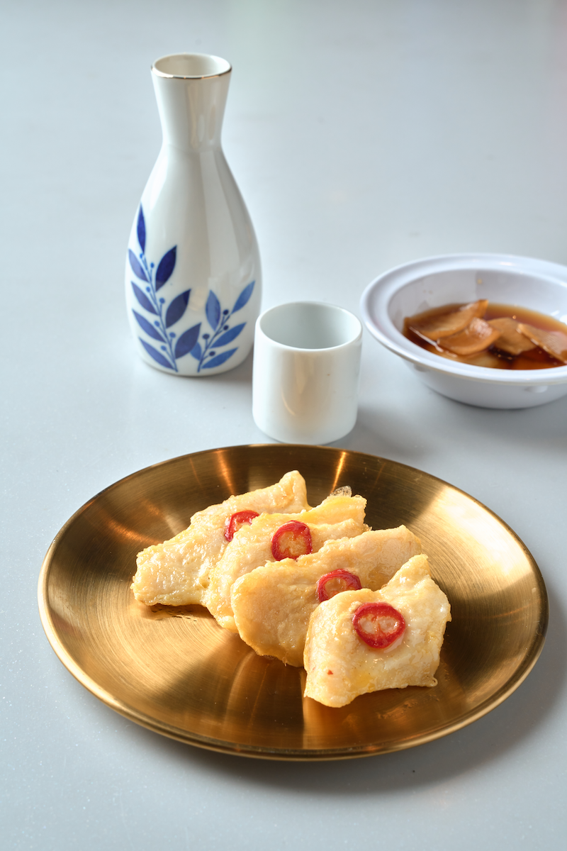 A gold plate with four pieces of fried fish topped with sliced red chili peppers on a white surface, with a white and blue vase, a small white cup, and a bowl of brown sauce in the background.