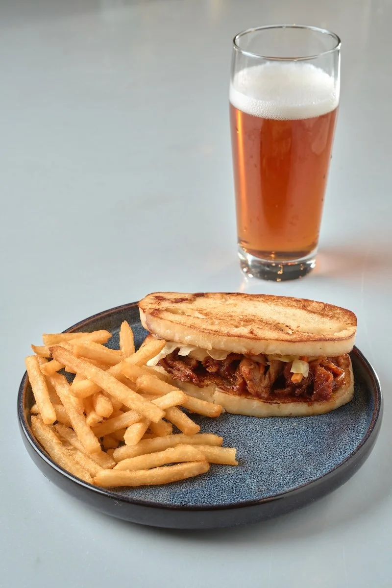 A plate with French fries and a sandwich bun filled with shredded meat, topped with grilled sandwich bread, served with a glass of beer.
